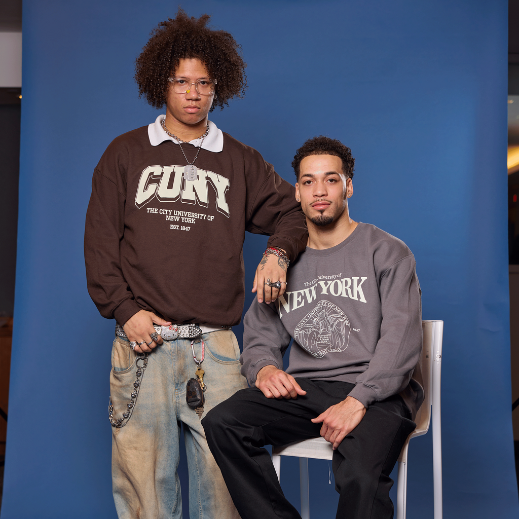 Two men posing against a blue backdrop, one sitting on a chair and the other standing.