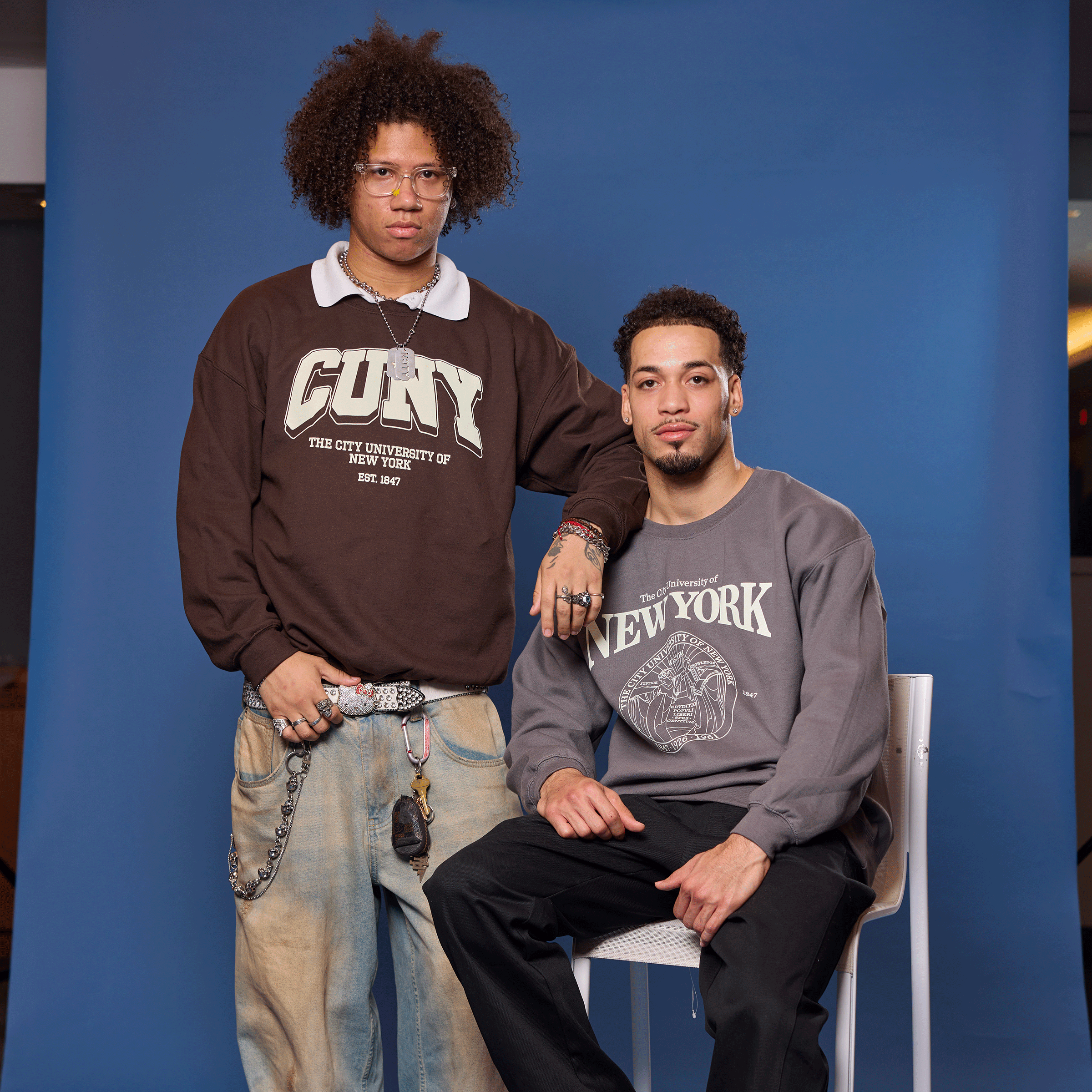 Two men posing against a blue backdrop, one sitting on a chair and the other standing.